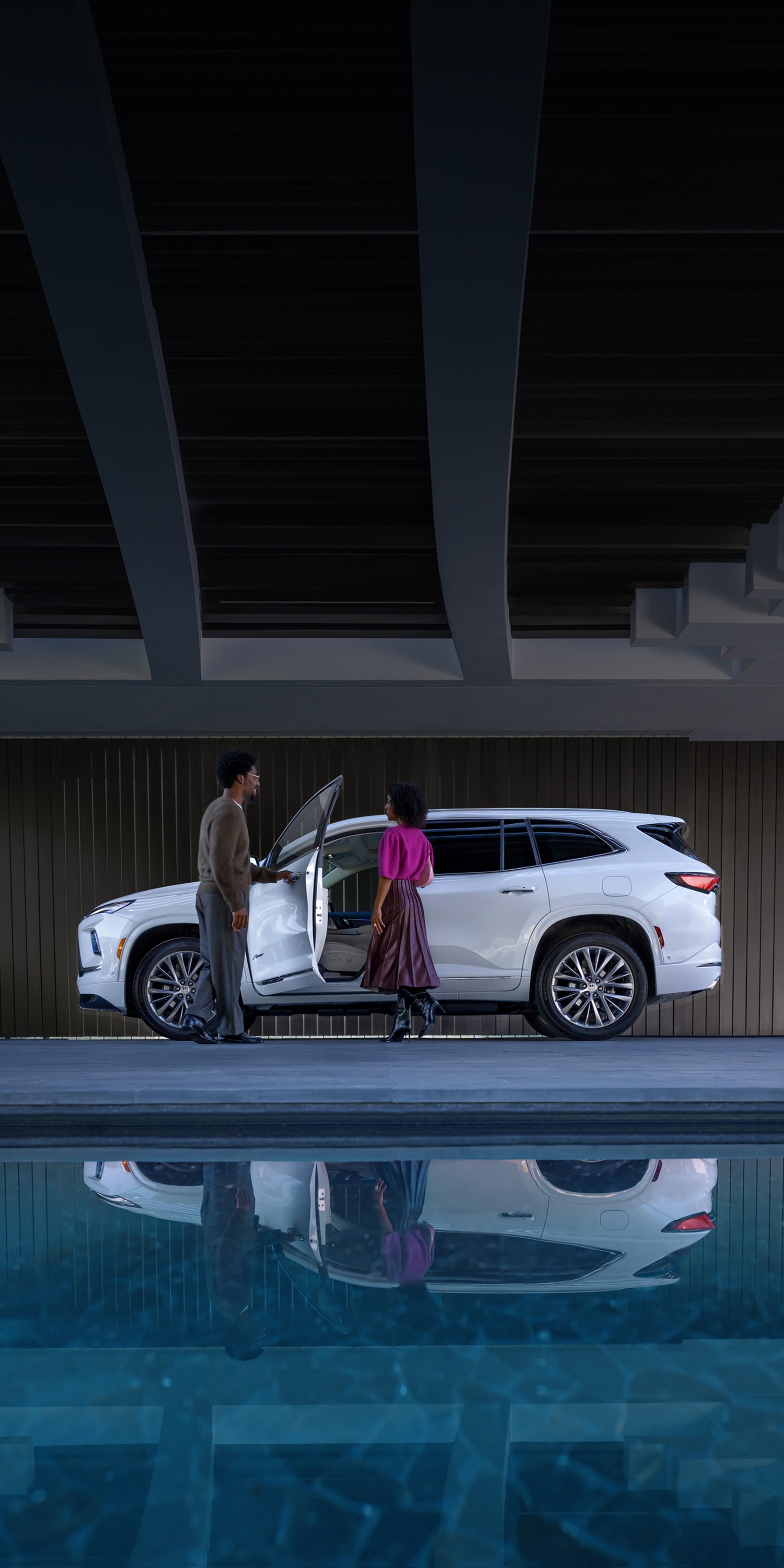 A Man and Woman Standing by a White Buick SUV with an Open Door, Reflected in a Pool of Water