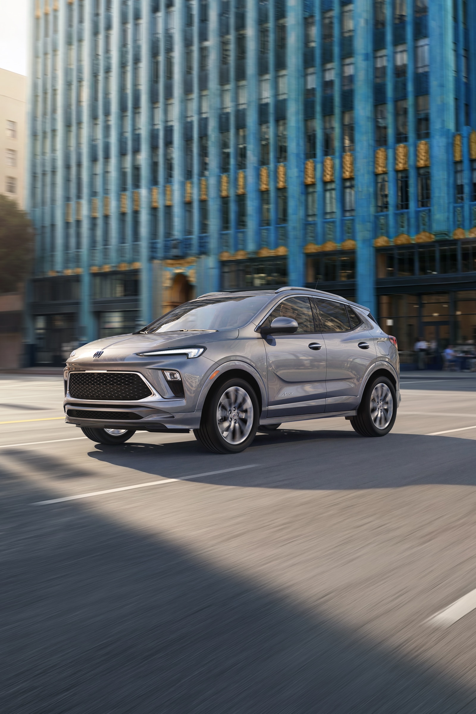A Silver Buick Encore Gx Suv Driving on a City Street, with a Modern Building in the Background