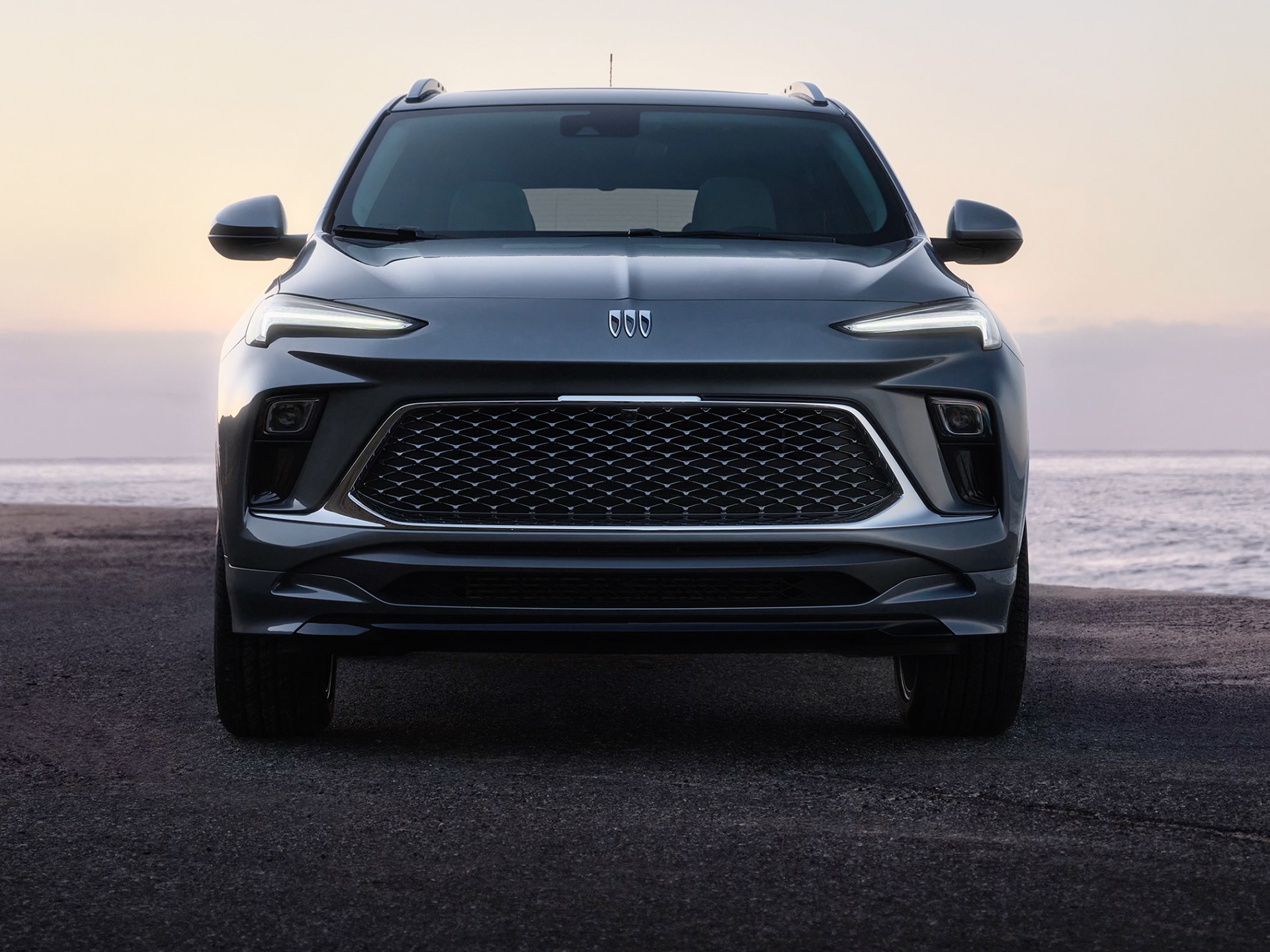 A Front View of a Dark Gray Buick Envista Suv Parked at Sunset by the Ocean, with Its Headlights On