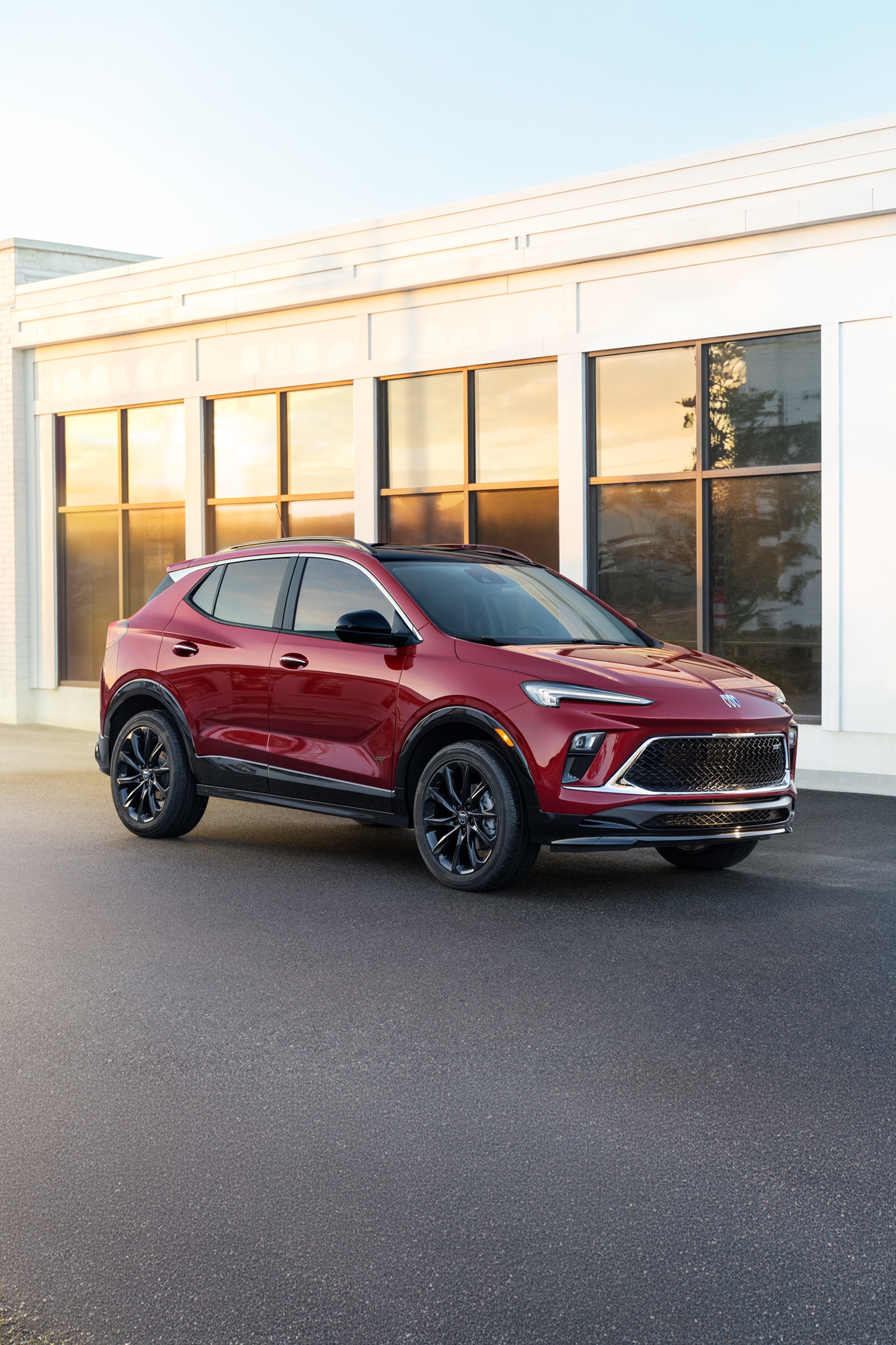 Front Three-Quarter View of a Dark Red Buick Encore GX Parked on Pavement with a Modern Building Reflecting Sunlight in the Background