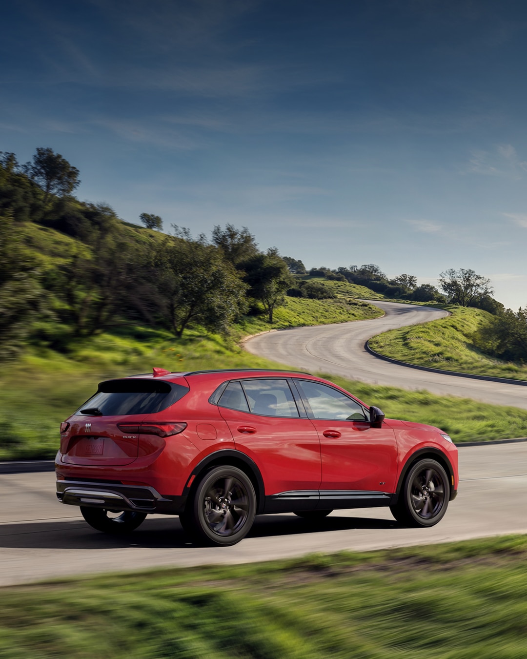 The Rear View of a Red Buick Envision SUV Driving on a Winding Road with Green Foliage