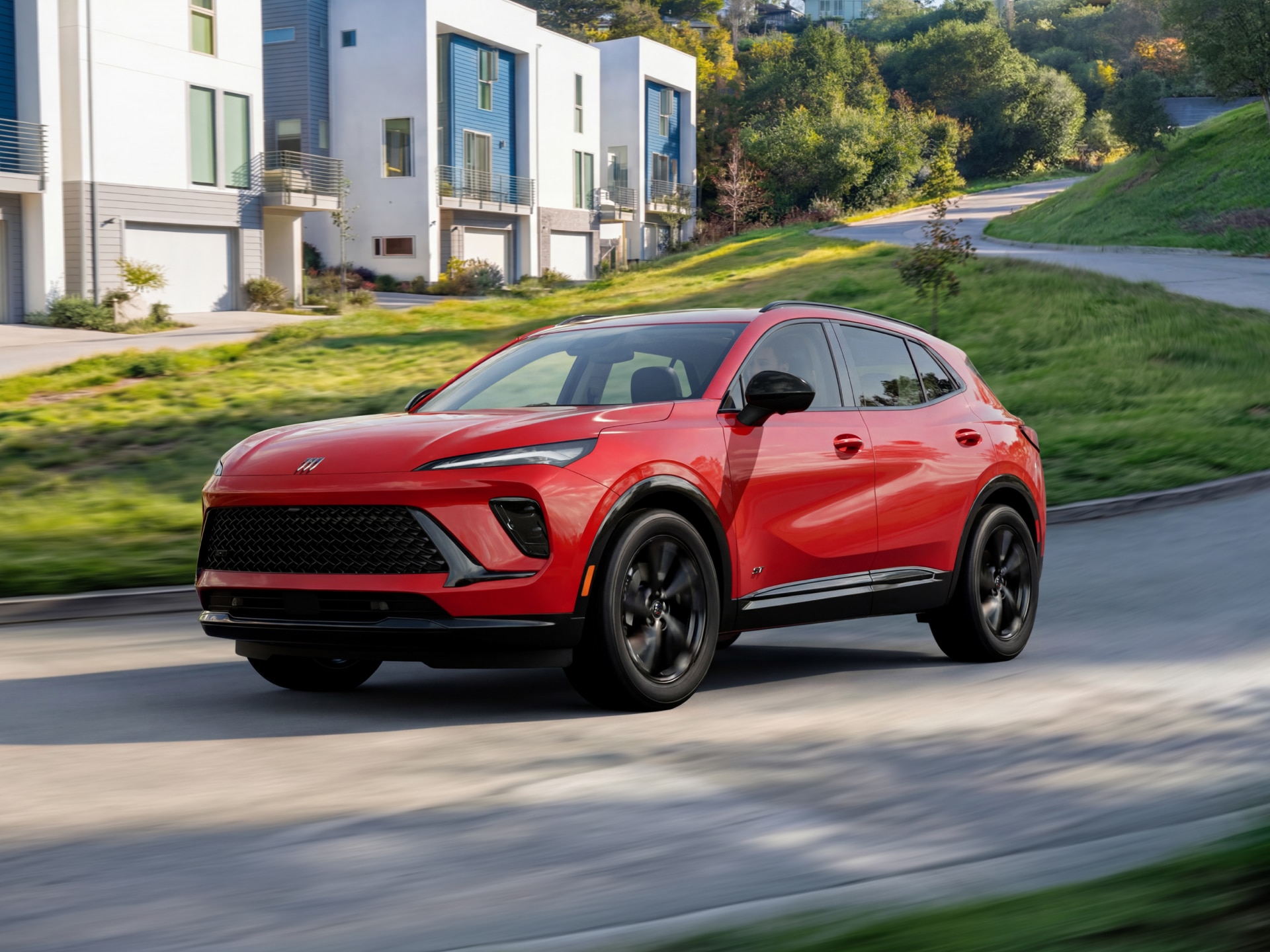 A Red 2027 Buick Envision Driving on a Paved Road with Modern Townhouses in the Background