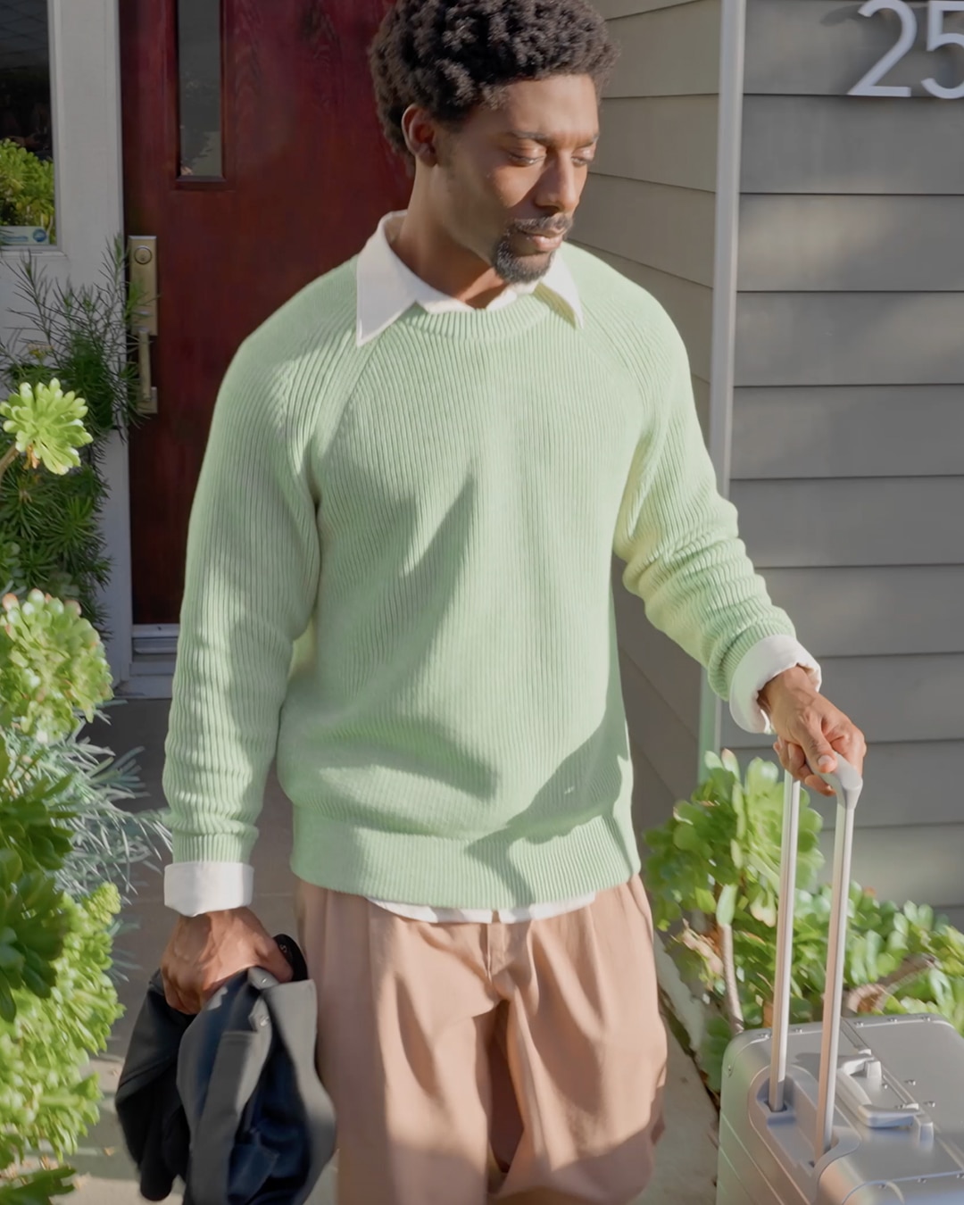 A Man in a Light Green Sweater and Brown Pants Walks Away from a House, Pulling a Silver Suitcase and Holding a Dark Jacket