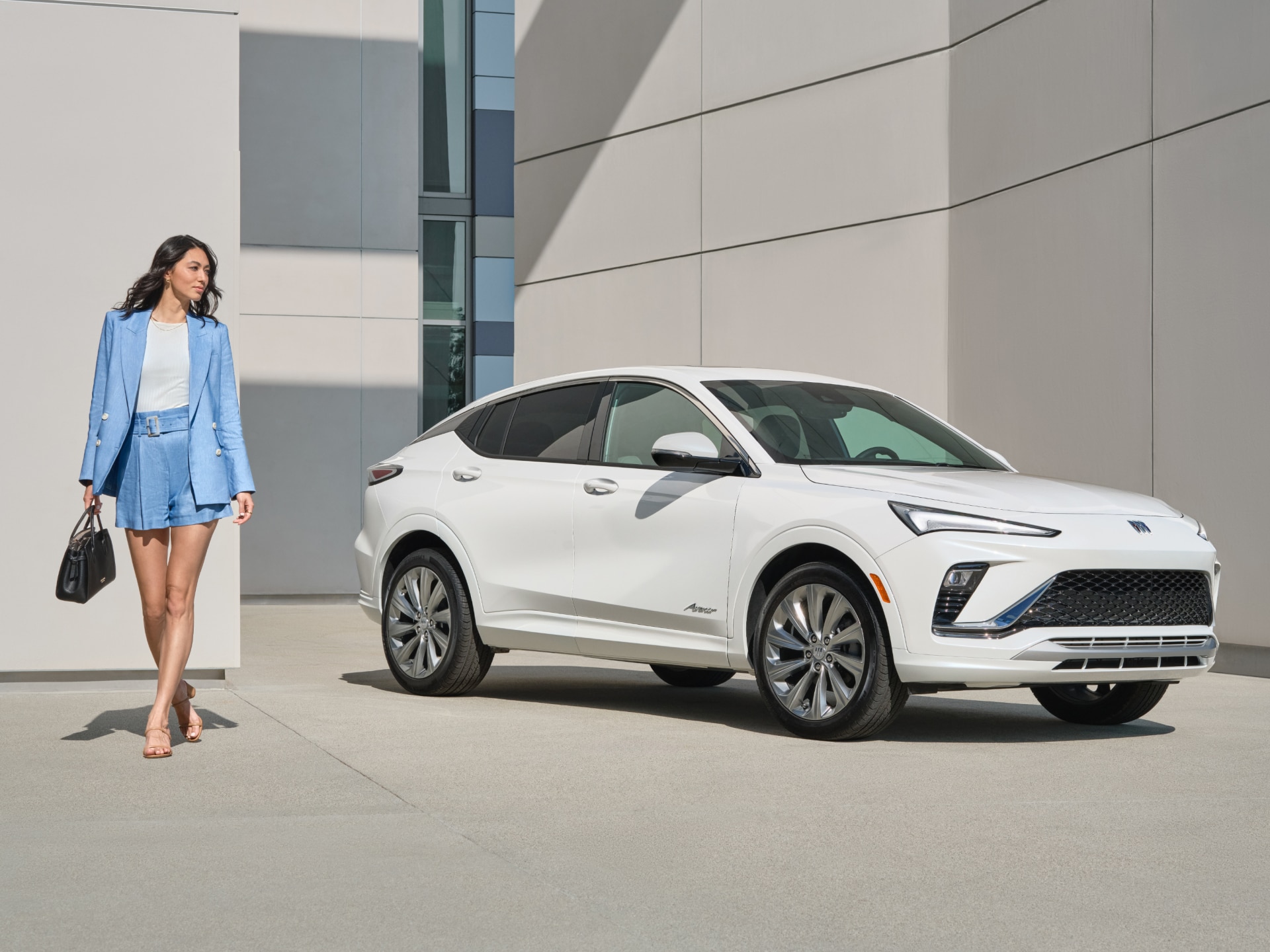 A Woman in a Blue Suit Walking Past a White Buick Envista Avenir Parked in Front of a Modern Building