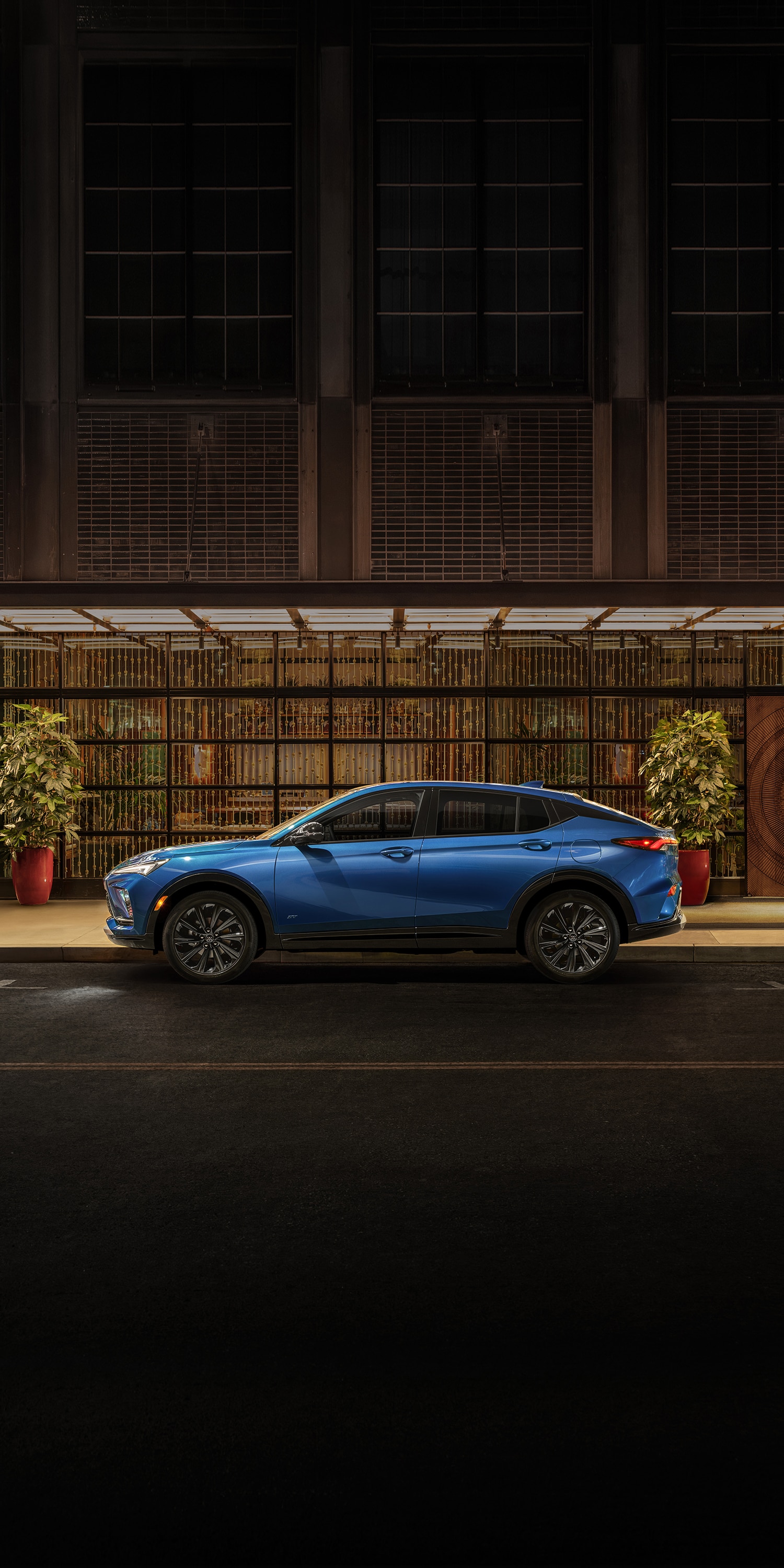 A Blue Buick Envista ST Parked on a City Street At Night with a Modern Building and Lush Green Plants