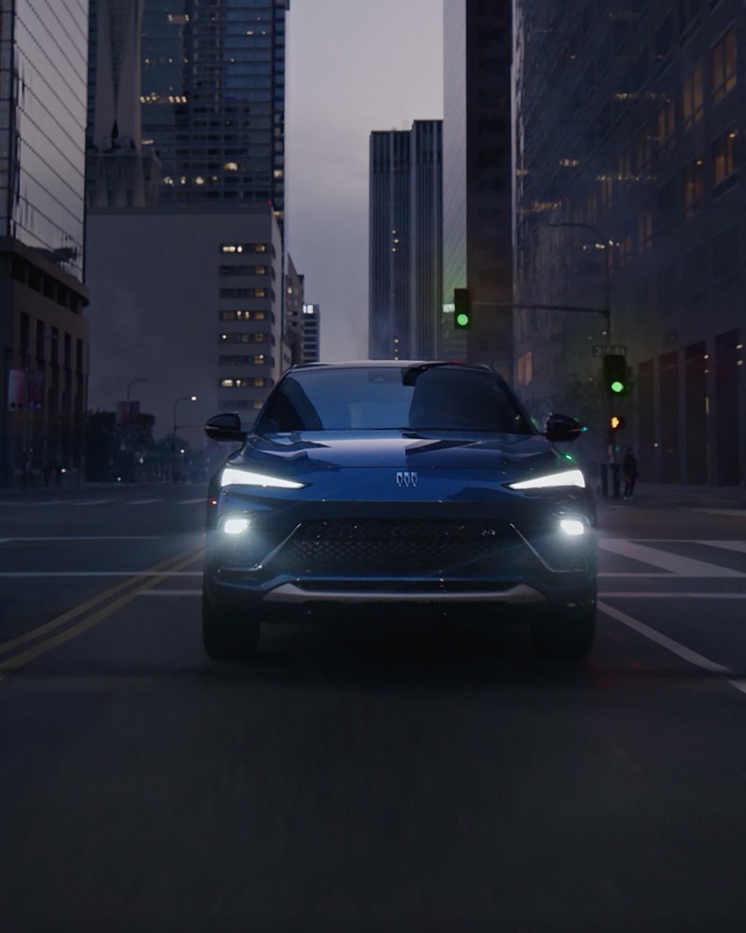 A Blue Buick SUV Driving Down a City Street at Dusk with Headlights Illuminated