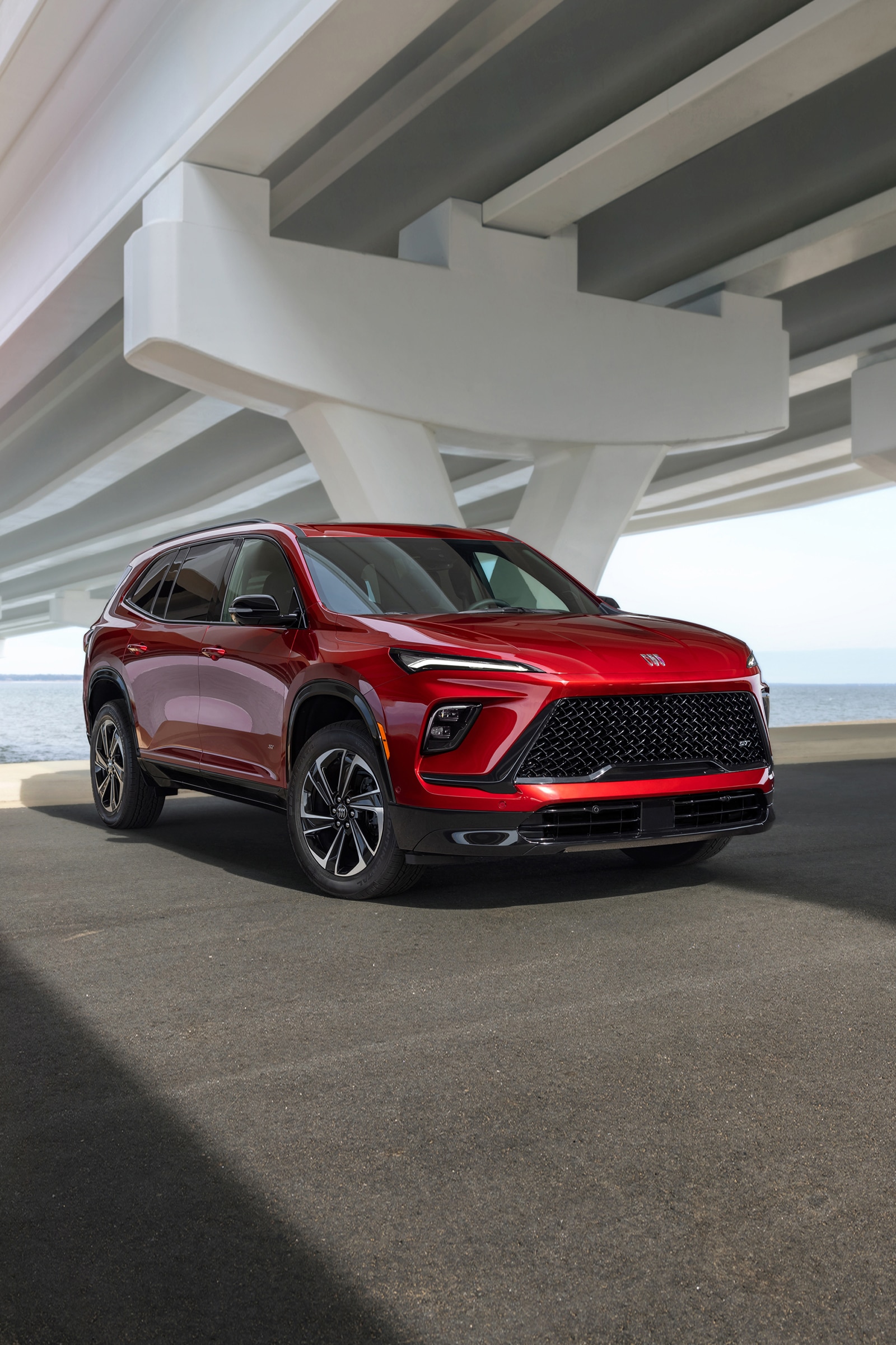 Red Buick Enclave SUV from a Front Three-Quarter Angle with a Bold Grille Under a Bridge and Water