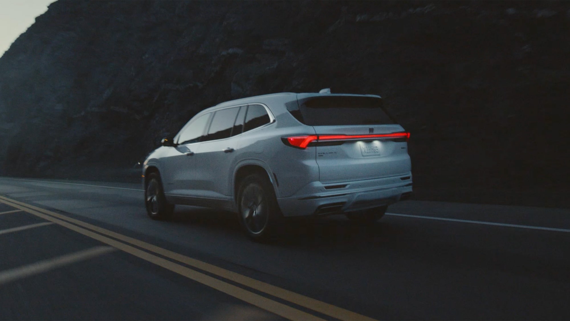 Three-Quarters Rear View of a 2026 White Buick Enclave Driving Down the Road Next to Mountains and a Body of Water