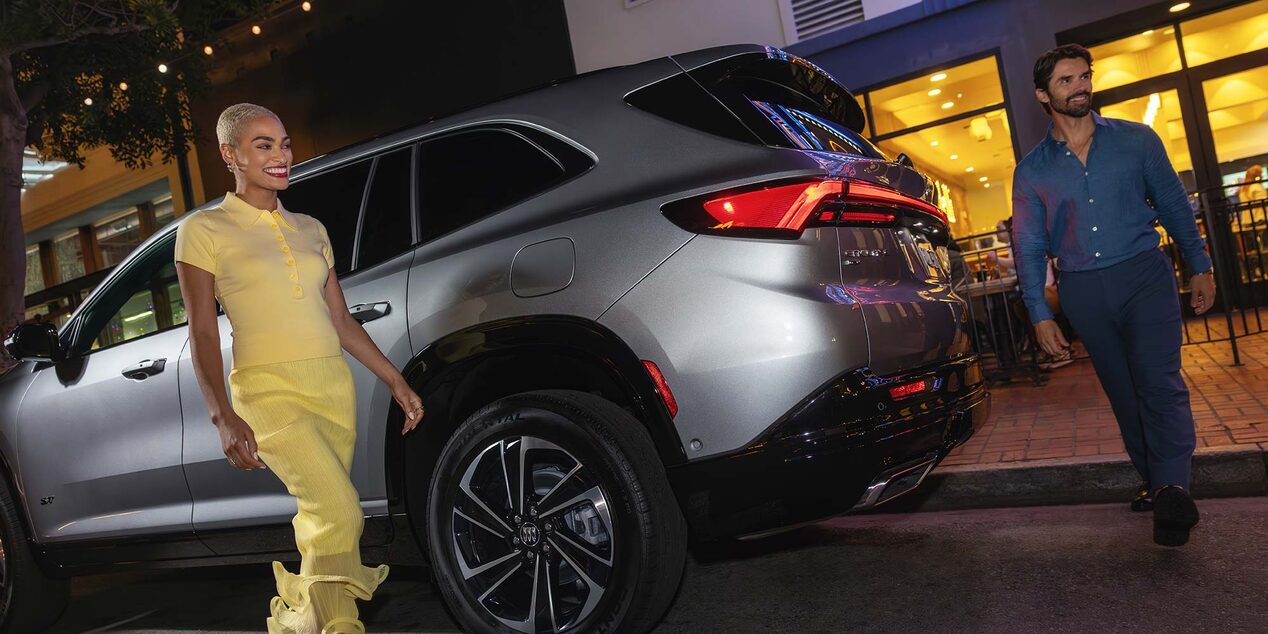 A Man and Woman Walking by a Gray Buick Enclave SUV on a City Street at Night