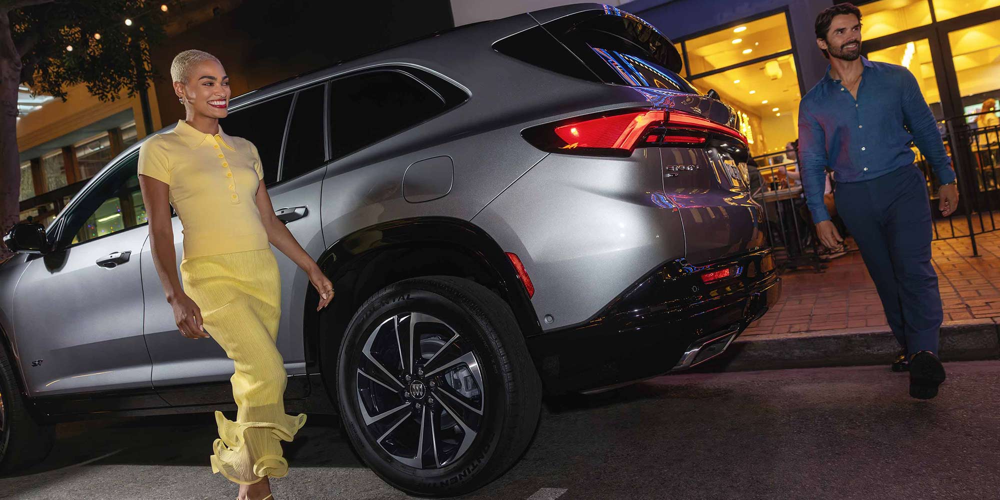 A Man and Woman Walking by a Gray Buick Enclave SUV on a City Street at Night