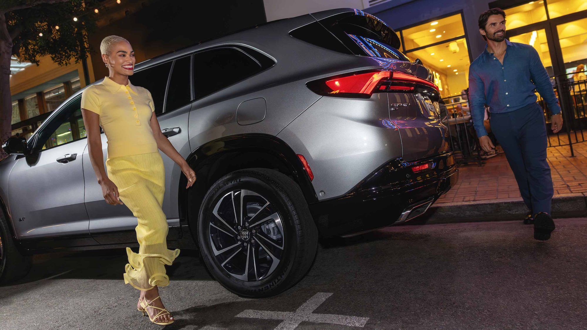 A Man and Woman Walking by a Gray Buick Enclave SUV on a City Street at Night